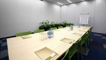 Sala de conferências com uma mesa comprida, cadeiras verdes, plantas em vasos, garrafas de água e um quadro branco.