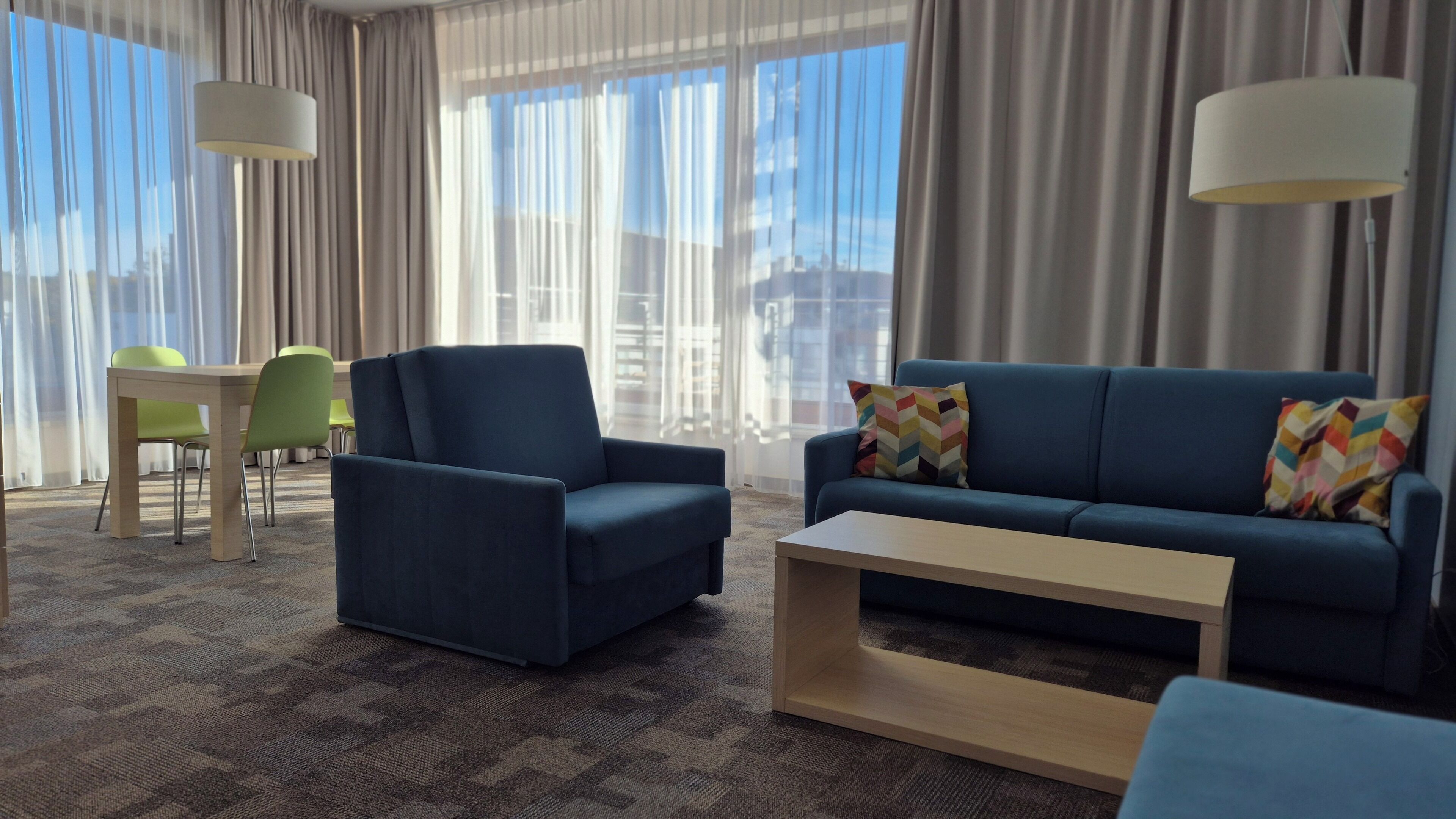 Una habitación de hotel moderna con un sofá azul, un sillón a juego, una mesa de centro de madera y una silla verde. La habitación cuenta con ventanas amplias con cortinas translúcidas, que dejan entrar abundante luz natural.