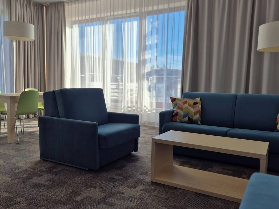 Una habitación de hotel moderna con un sofá azul, un sillón a juego, una mesa de centro de madera y una silla verde. La habitación cuenta con ventanas amplias con cortinas translúcidas, que dejan entrar abundante luz natural.