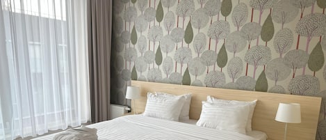 A hotel room with a large bed, white linens, a headboard with a leaf pattern wallpaper, two bedside tables with lamps, and a window with sheer curtains.