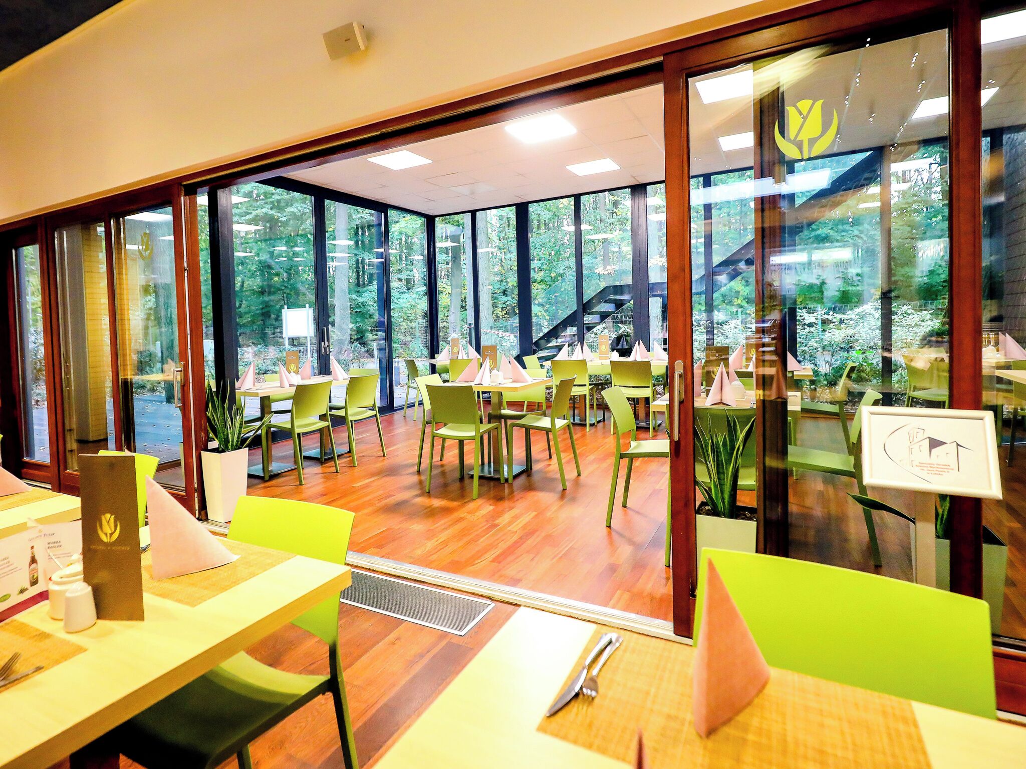 A dining area with tables and chairs, large windows, and a view of greenery outside.