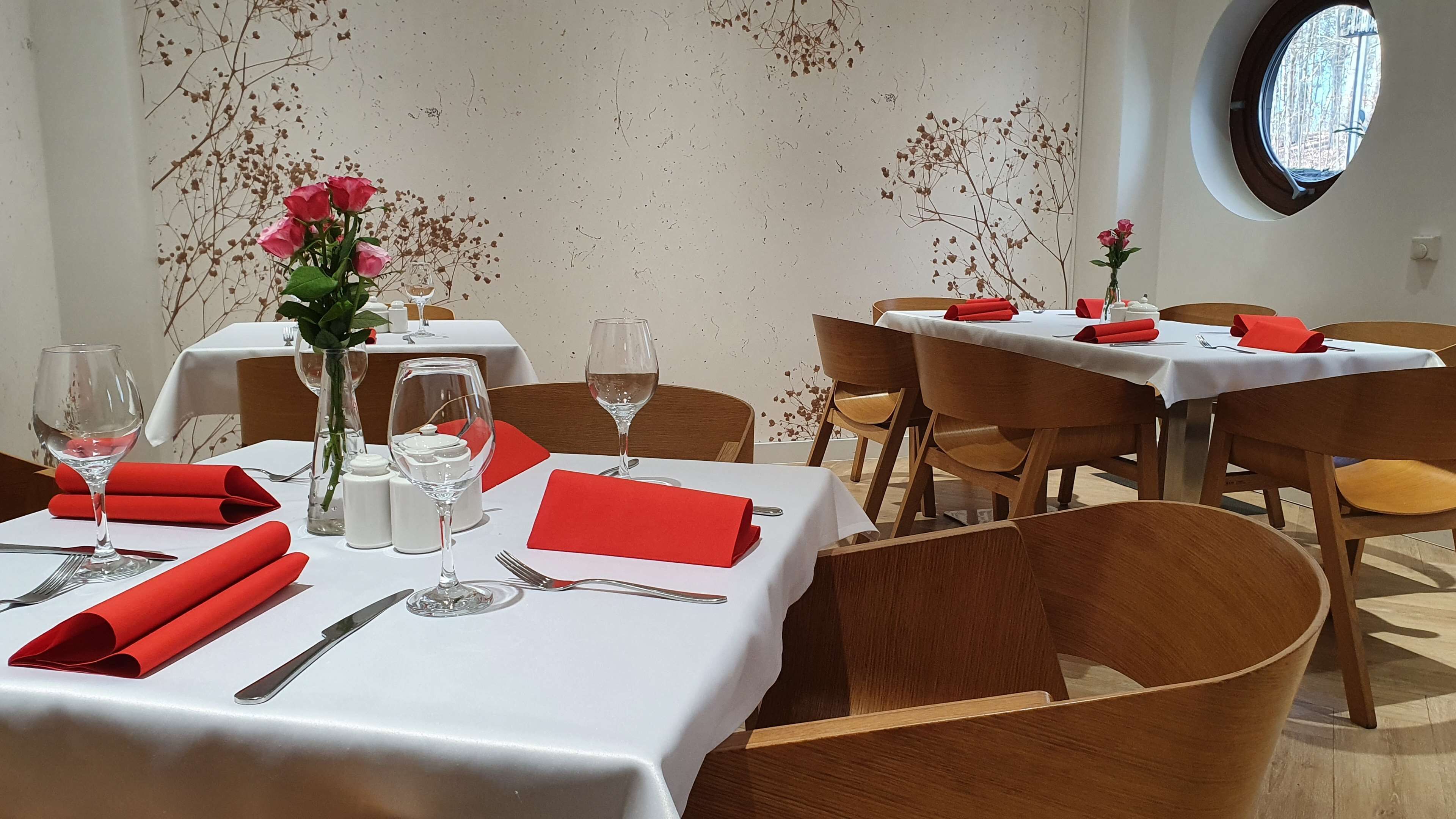 A restaurant interior with round tables, white tablecloths, red napkins, and a vase with pink flowers.
