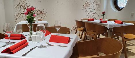 A restaurant interior with round tables, white tablecloths, red napkins, and a vase with pink flowers.