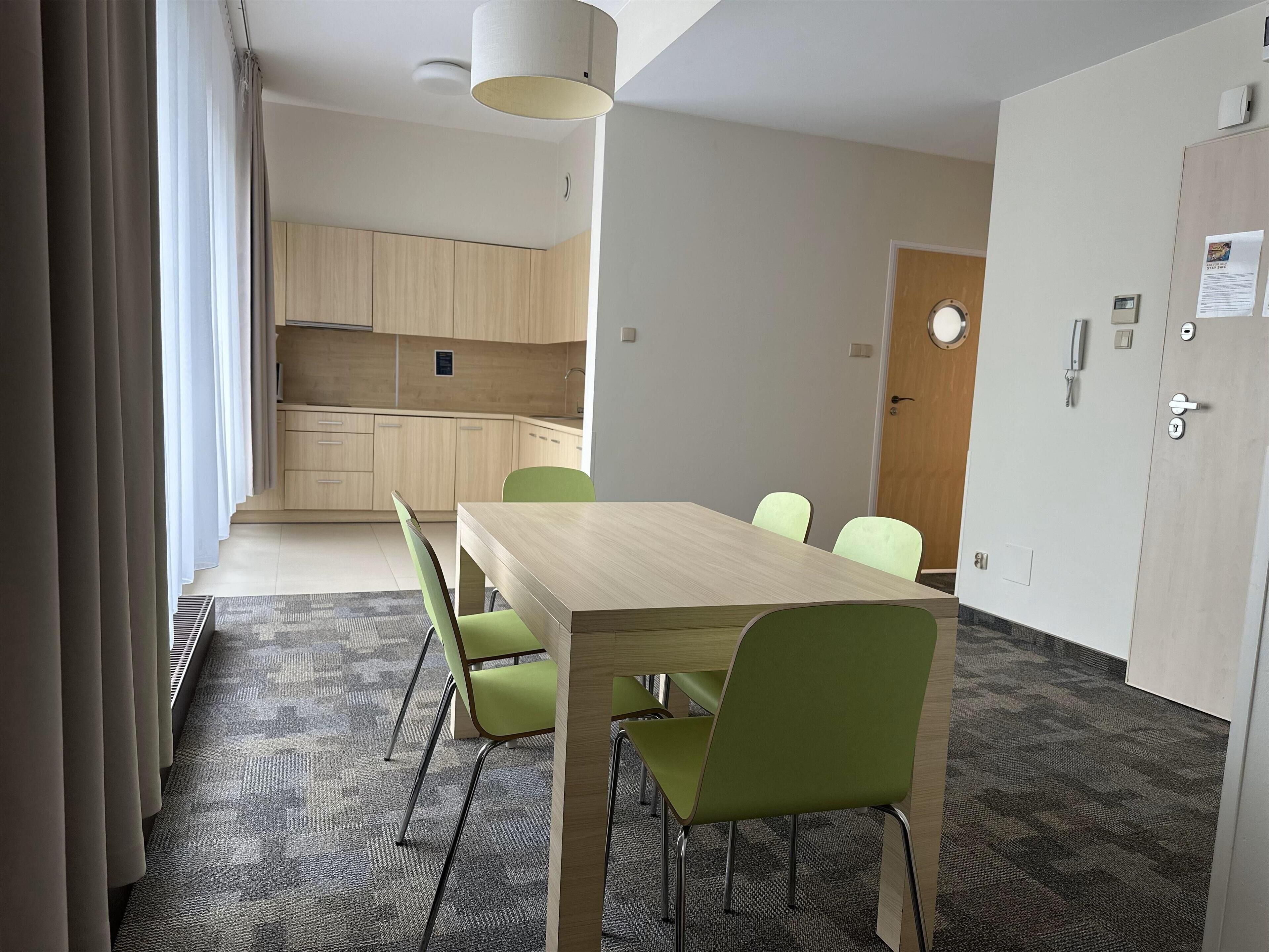 A modern hotel room with a wooden dining table and green chairs, a kitchenette, and a door with a keycard lock.