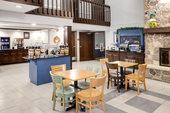 A modern hotel lobby with a central island, wooden tables and chairs, a stone fireplace, and a staircase.