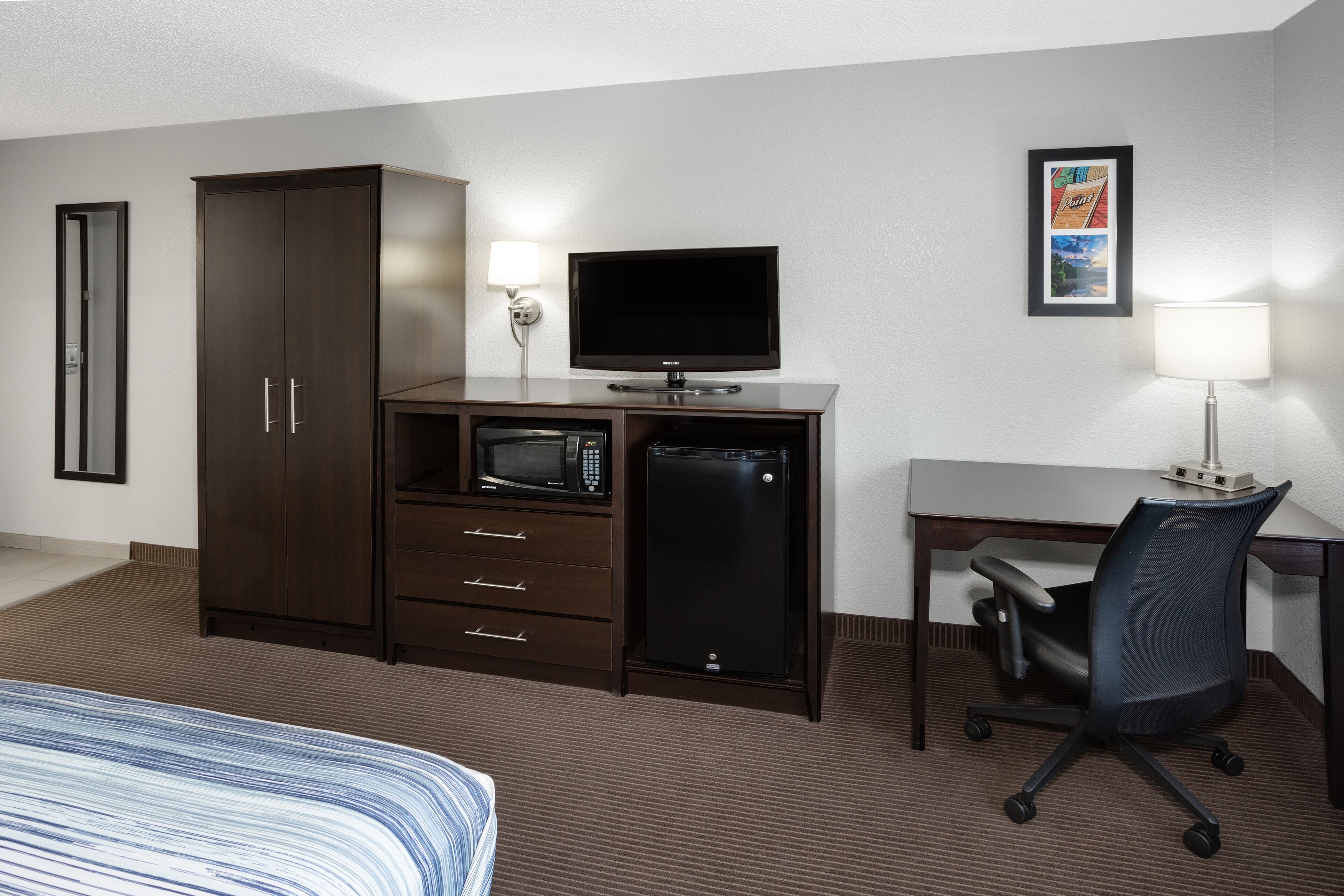 Habitación de hotel con cama, escritorio, silla, armario, televisor, microondas, refrigerador y dos lámparas de pared.