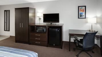 Habitación de hotel con cama, escritorio, silla, armario, televisor, microondas, refrigerador y dos lámparas de pared.