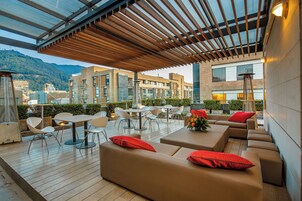 Un área de estar al aire libre con mesas y sillas, una pérgola de madera y vistas a edificios al fondo.