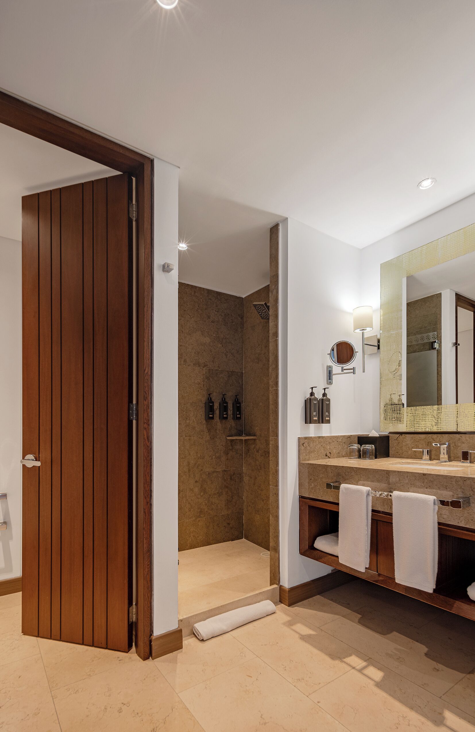 A modern bathroom with a wooden door, a large mirror, a shower area, and a vanity with multiple sinks and toiletries.