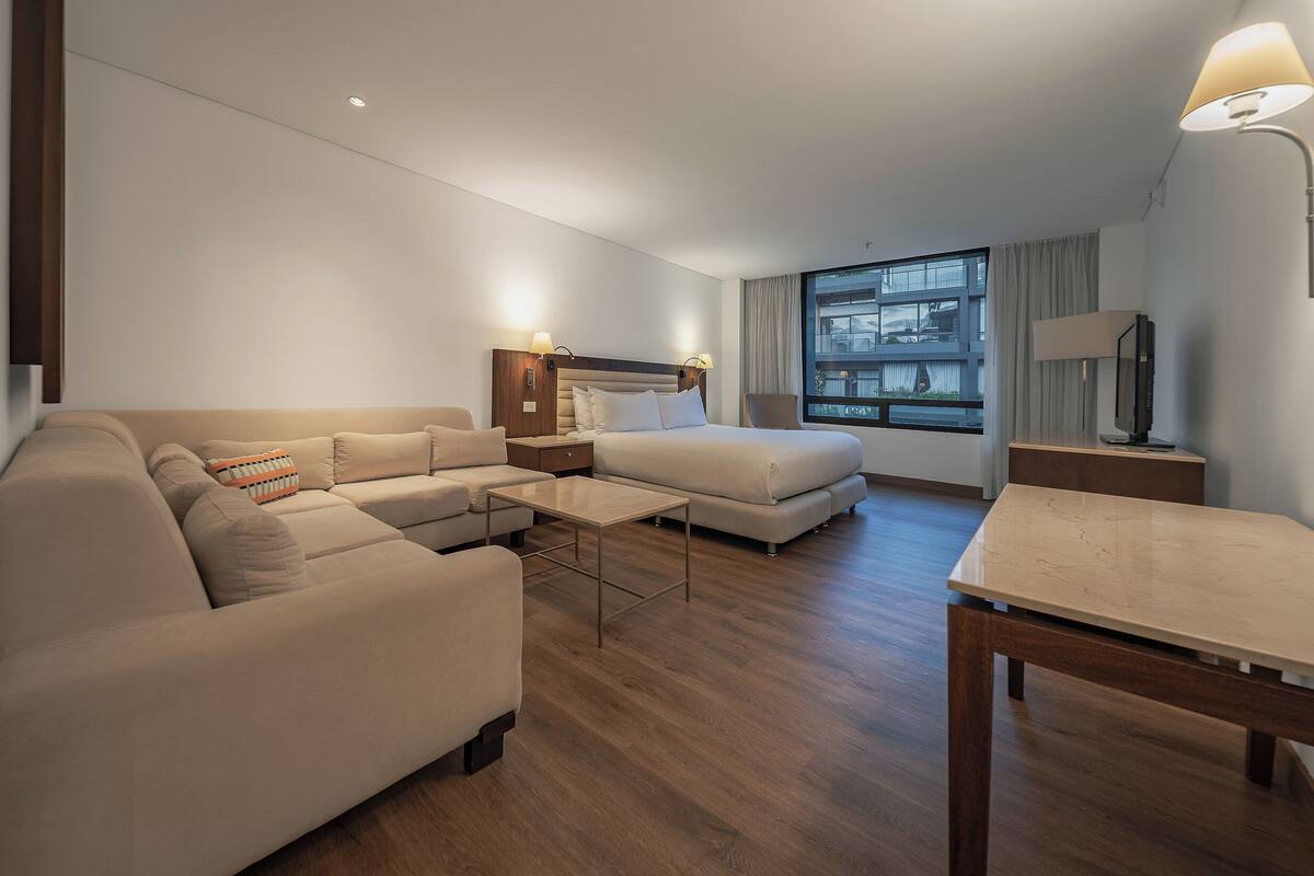 A modern hotel room with a sofa, a small table, a TV, and a window with curtains.