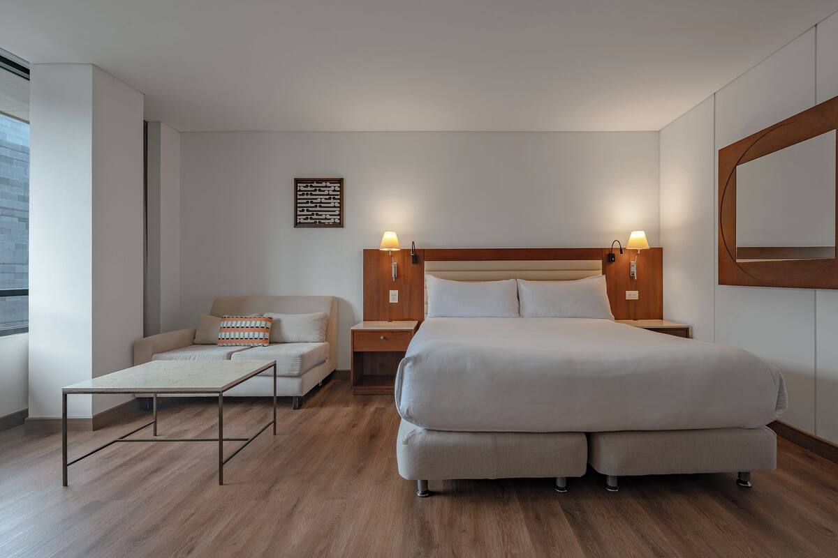 A modern hotel room with a large bed, a sofa, a small table, and a nightstand with lamps.