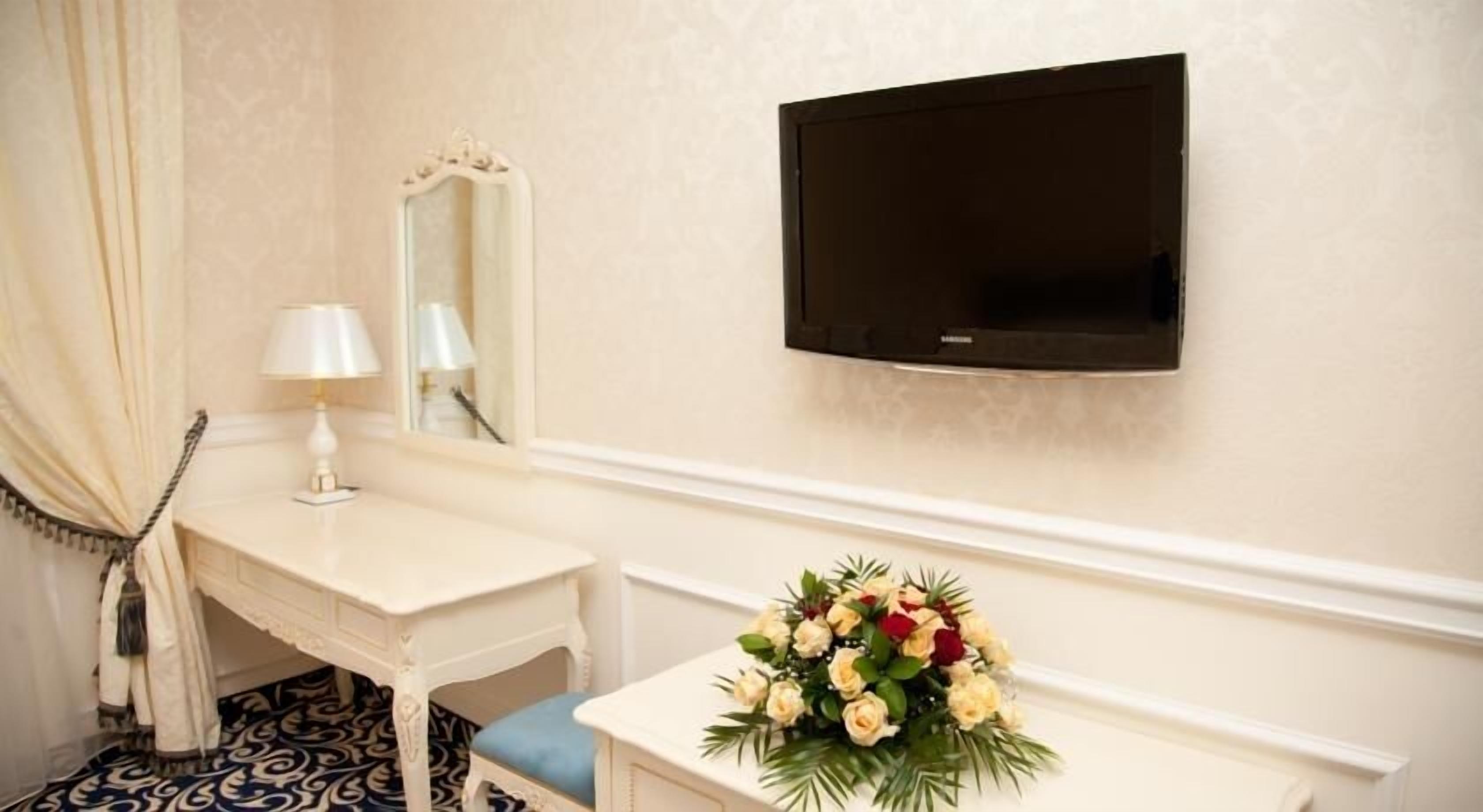 A hotel room with a white vanity, a flat-screen TV mounted on the wall, a mirror above the vanity, a telephone, and a floral arrangement on the vanity.