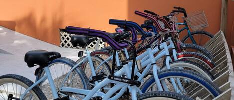 A row of bicycles parked under a covered area with a white roof, adjacent to a building with an orange wall and a stone facade.