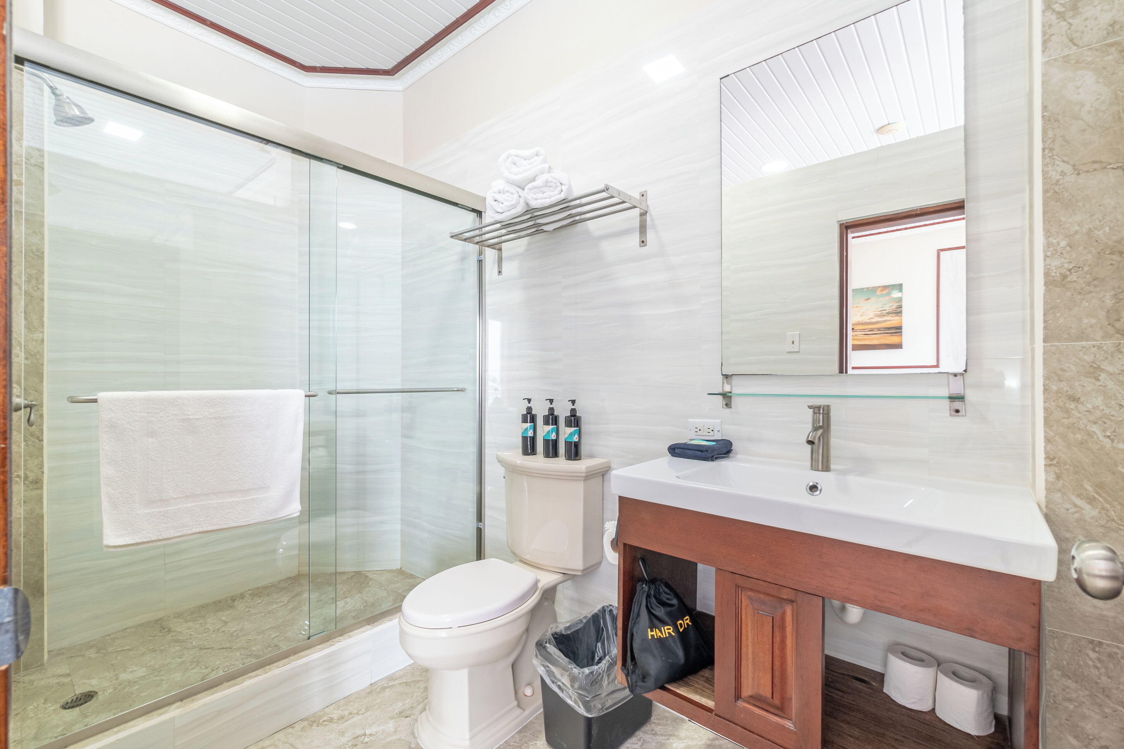 Ein modernes Badezimmer mit Duschkabine aus Glas, Toilette, Waschbecken mit Spiegel und einem Handtuchhalter mit Flaschen.