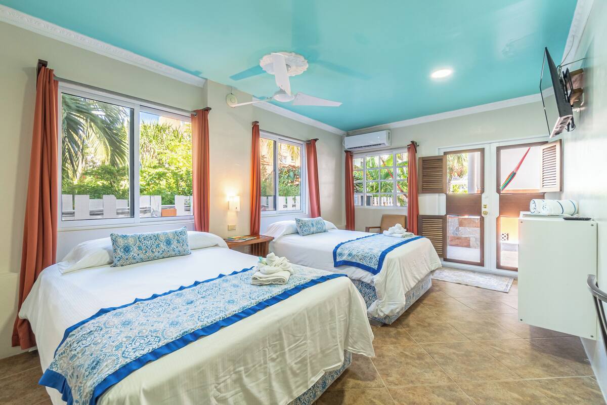 A hotel room with two beds, a ceiling fan, a TV, and a view of greenery outside.