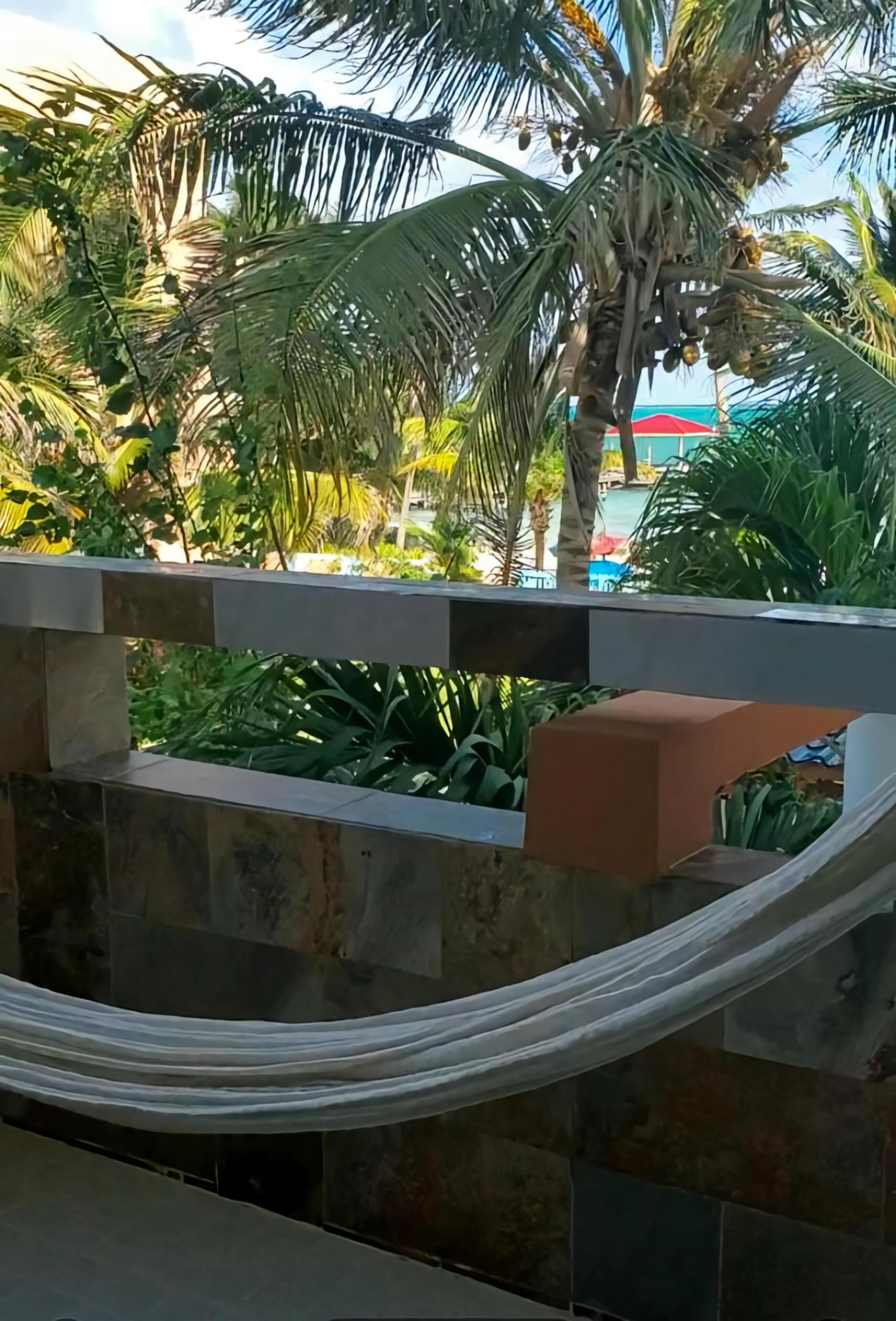 Una hamaca atada entre un muro de piedra y una barandilla, con vista a una zona de piscina con palmeras y un paraguas rojo.
