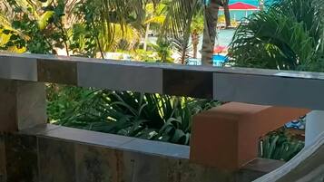 Una hamaca atada entre un muro de piedra y una barandilla, con vista a una zona de piscina con palmeras y un paraguas rojo.