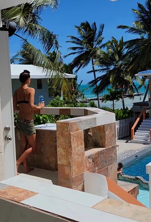 Zona de piscina con un banco de piedra, una persona sosteniendo una botella de agua y vista al océano con palmeras.