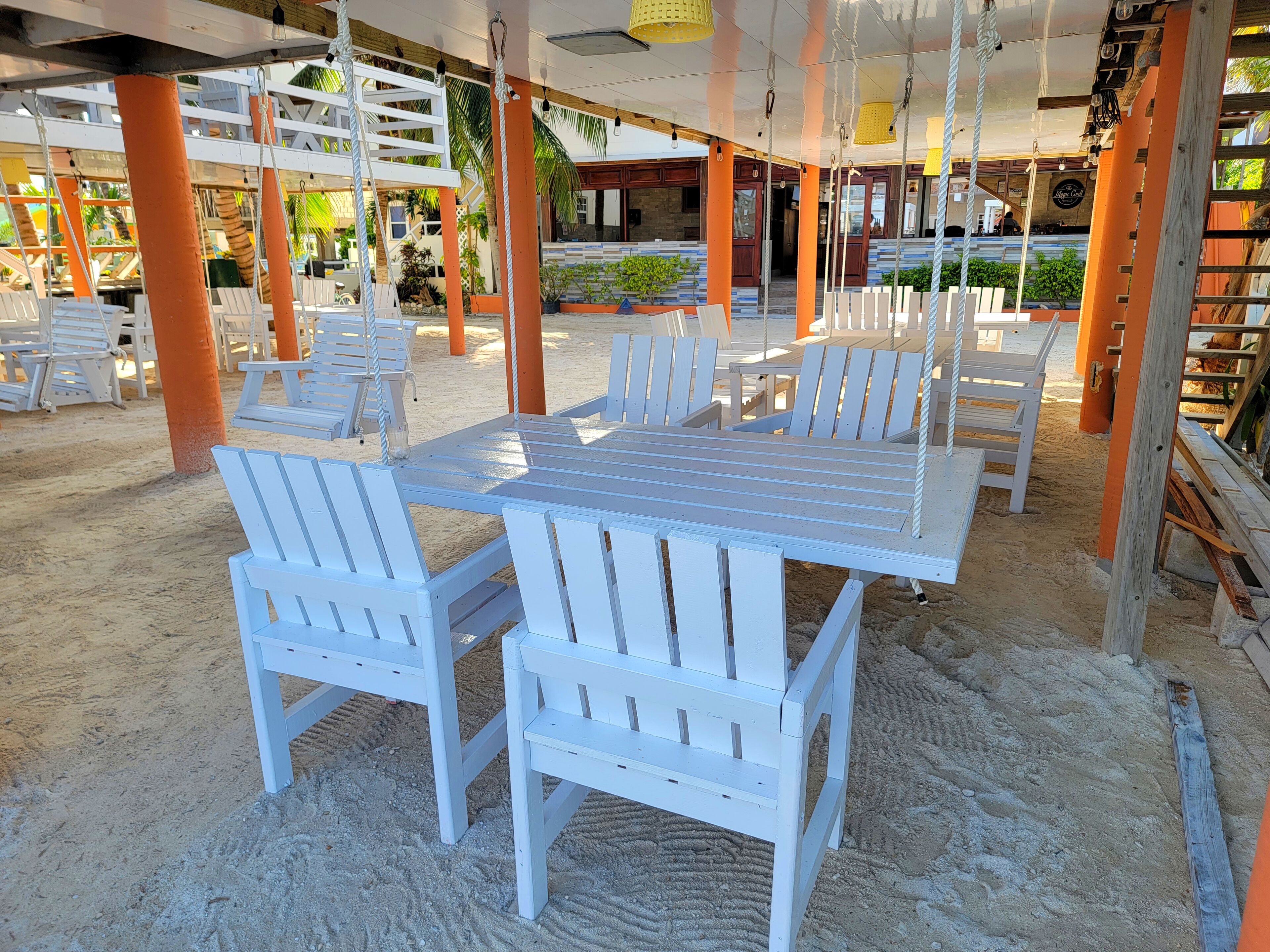 Un restaurant situé en bord de mer, avec des tables et des chaises en bois peintes en blanc, un espace de restauration couvert et une vue sur l’océan.