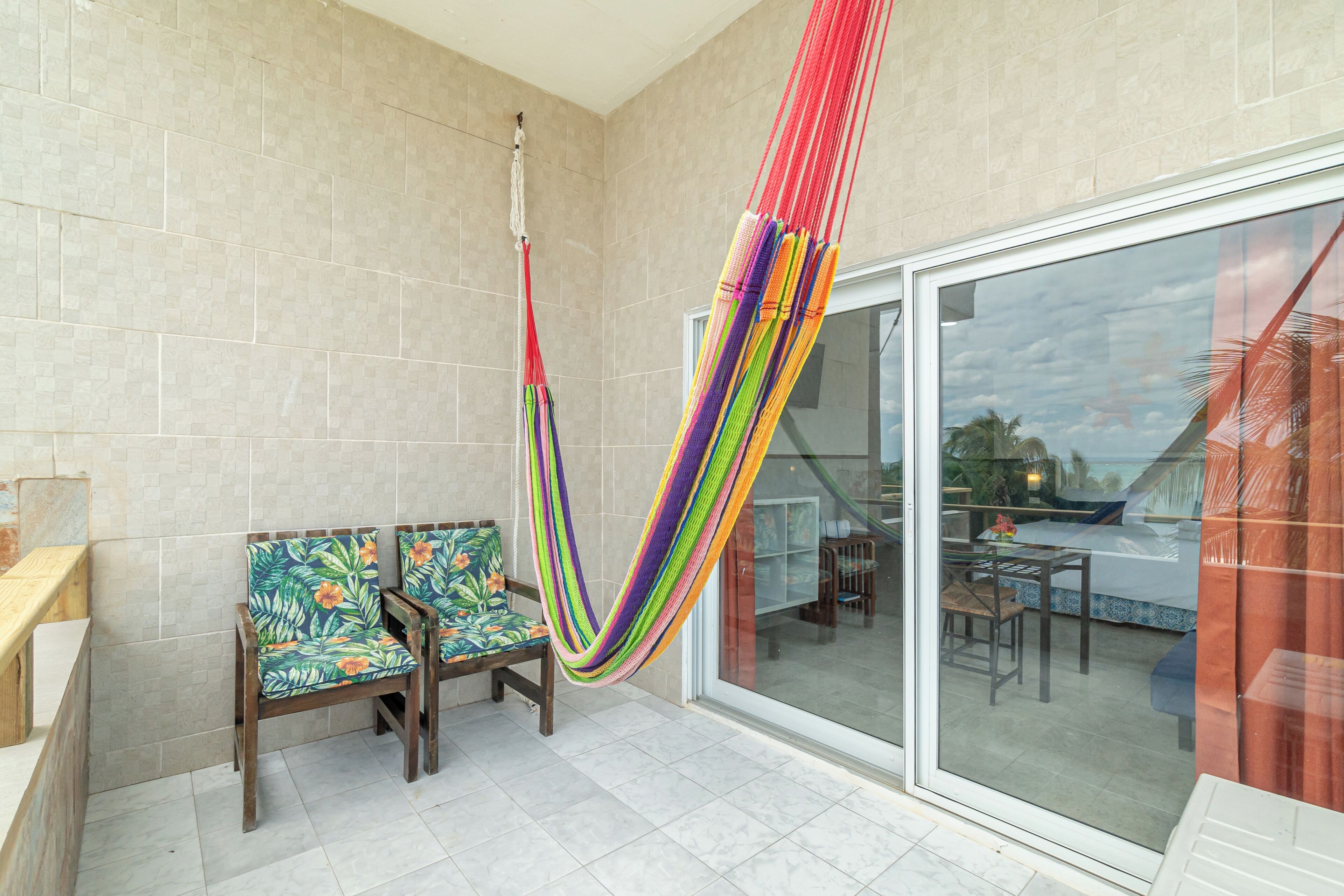 A hammock in a room with a glass door leading to a balcony, two wooden chairs with floral upholstery, and a wooden bench.