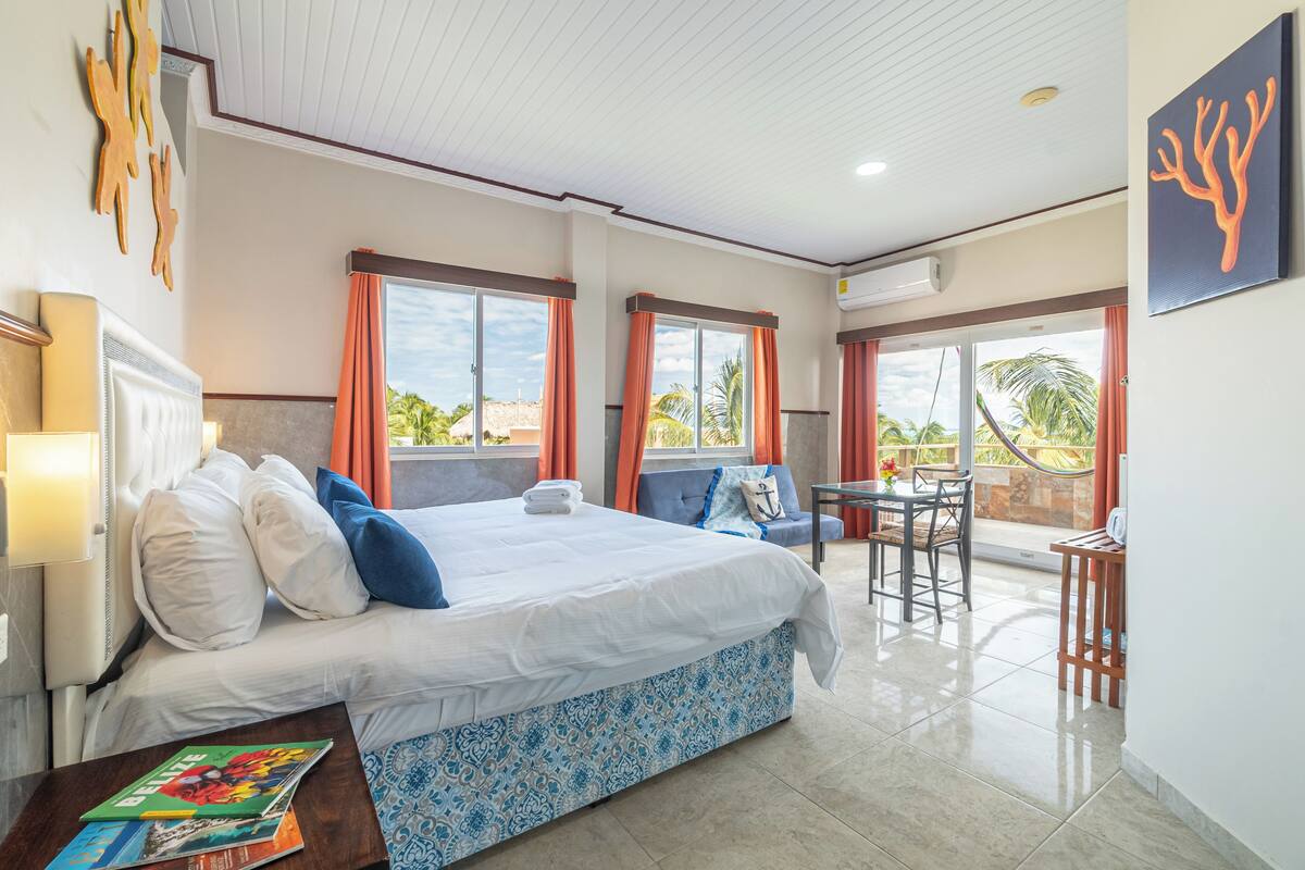 A hotel room with a bed, a small table, a chair, a balcony with a view of palm trees, and a ceiling fan.