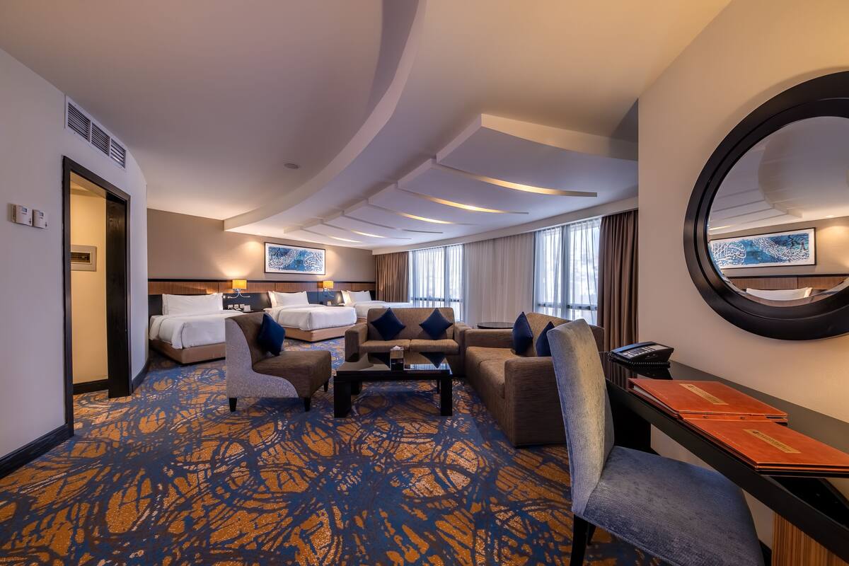 A hotel room with a bed, two sofas, a coffee table, a desk with a phone and folders, and a large mirror.