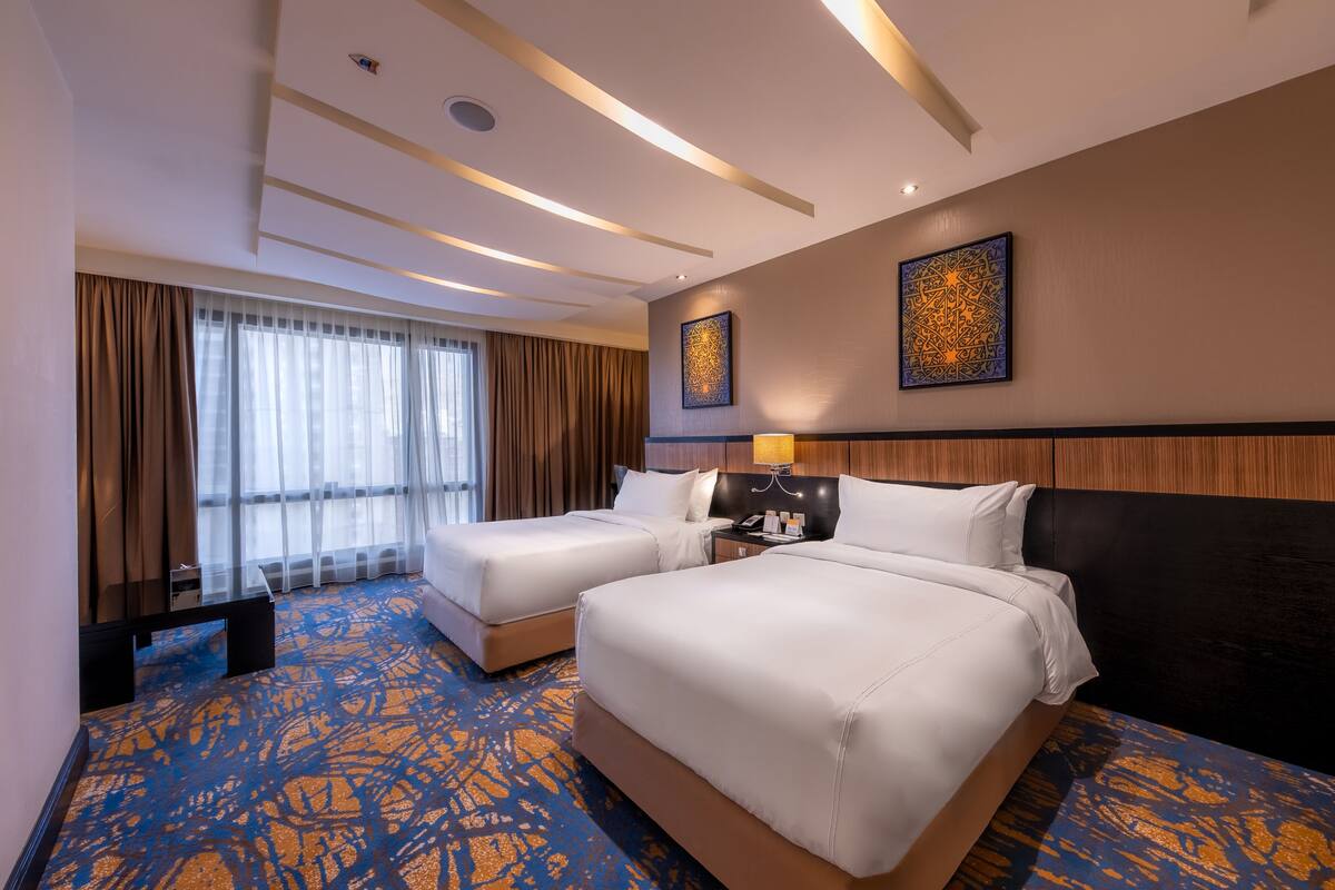 A hotel room with two beds, a large window with curtains, a wall-mounted artwork, and a patterned carpet.