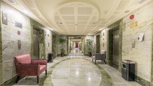 A hotel lobby with marble floors, a red sofa, a grey armchair, and two elevators.