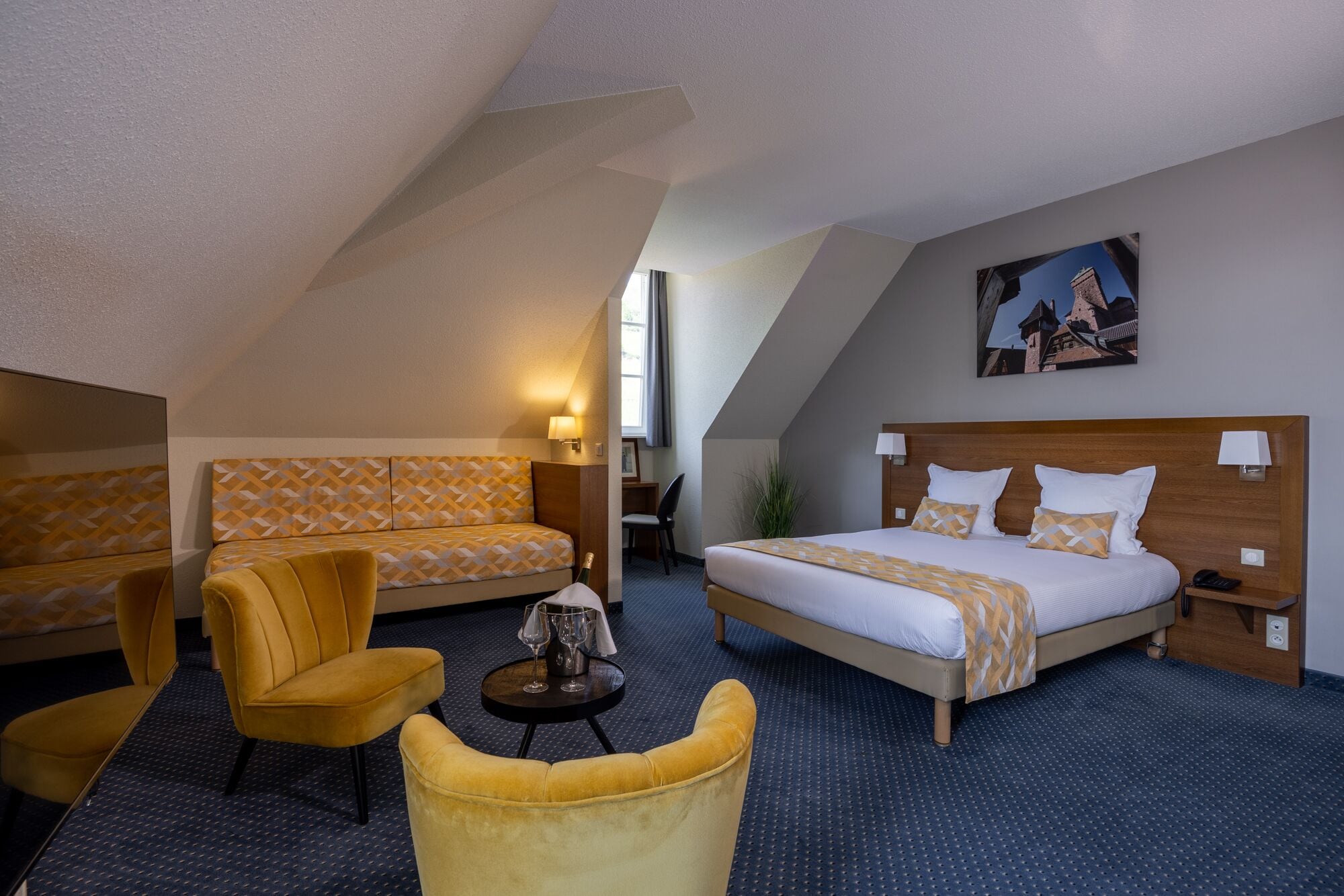 A hotel room with a large bed, a sofa, and a chair. There is a painting on the wall and a window with curtains.