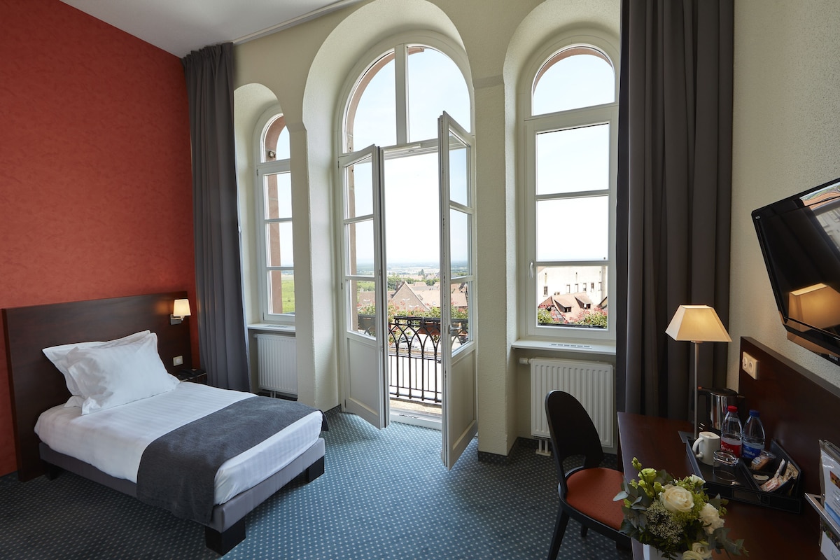 A hotel room with a bed, a desk with a chair, a TV, and large windows with a view.