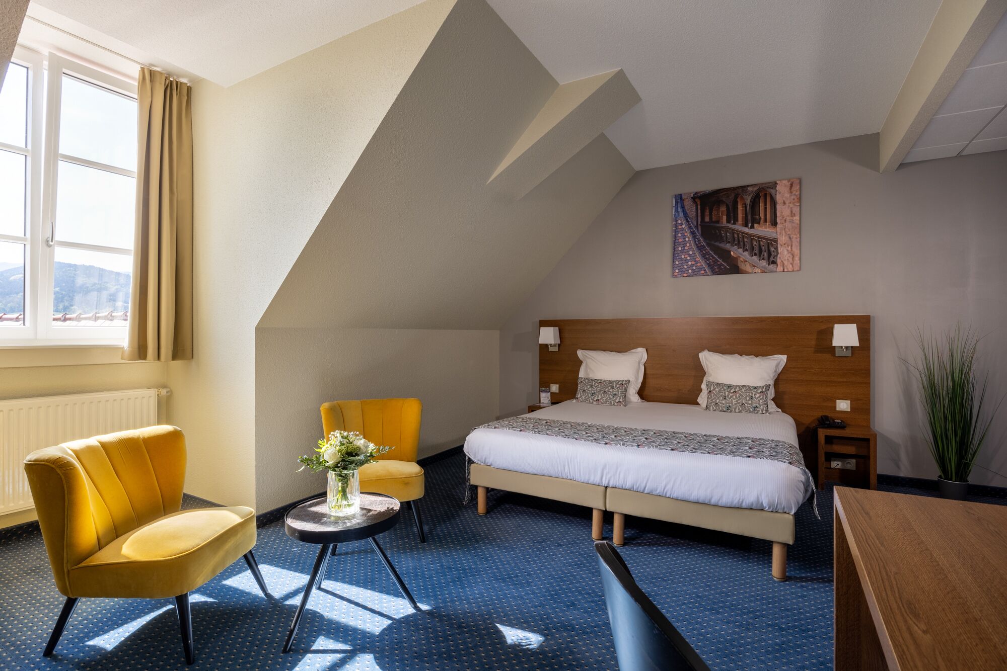 A hotel room with a bed, two chairs, a small table with a vase, a nightstand, and a painting on the wall.