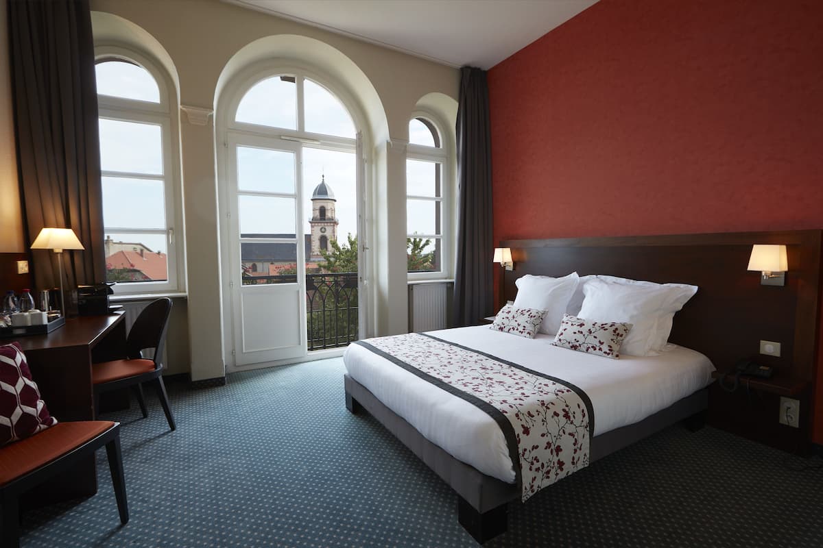 Ein Hotelzimmer mit einem großen Bett, einem Schreibtisch mit Stuhl und Blick auf einen Turm durch Bogenfenster.