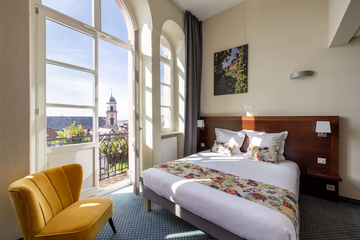 Ein Hotelzimmer mit einem großen Bett, einem gelben Sessel und Blick auf eine Stadtlandschaft durch große Fenster.