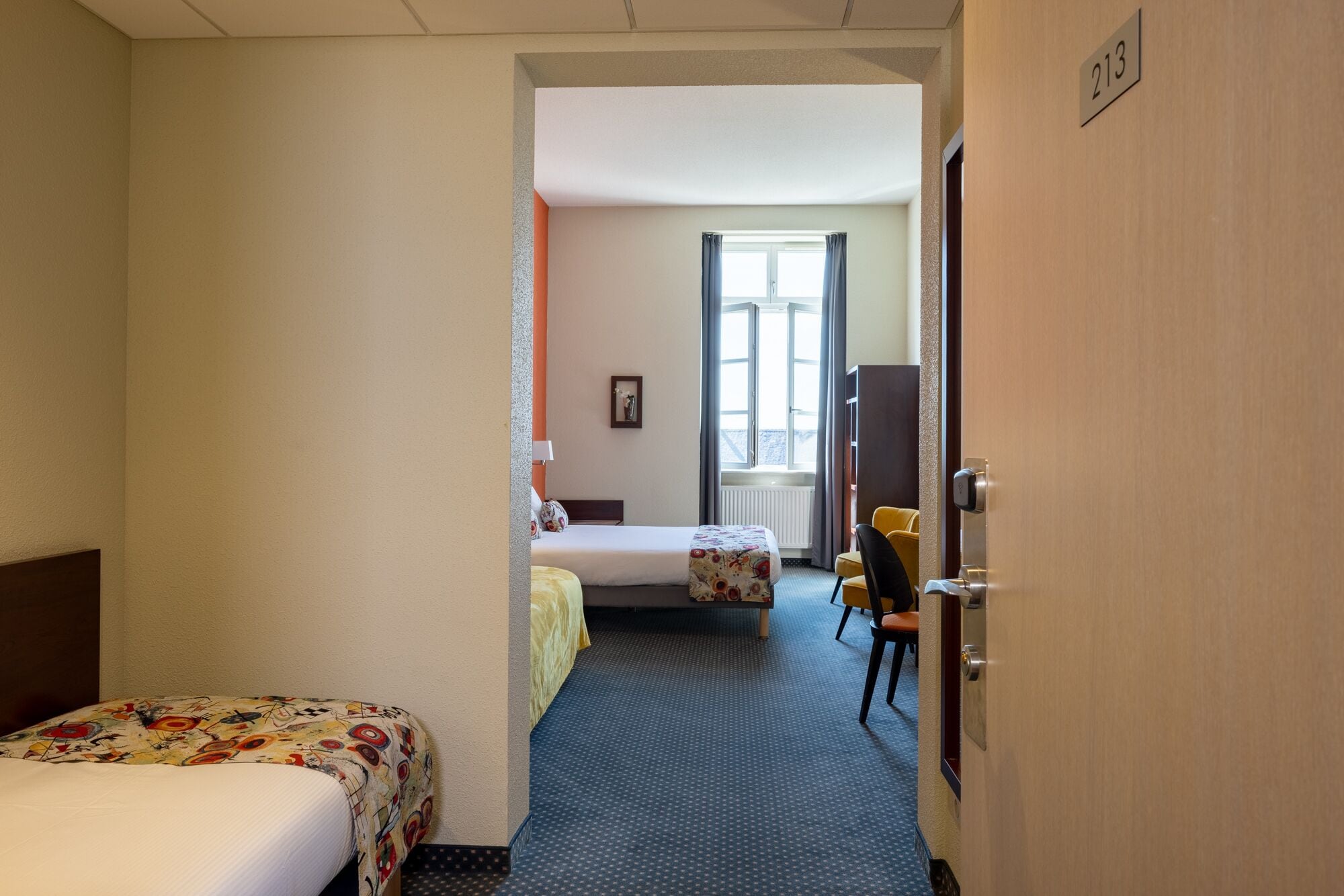 A hotel room with two beds, a desk, a chair, a window with curtains, and a door with a number sign.