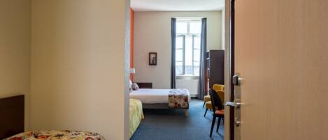 A hotel room with two beds, a desk, a chair, a window with curtains, and a door with a number sign.