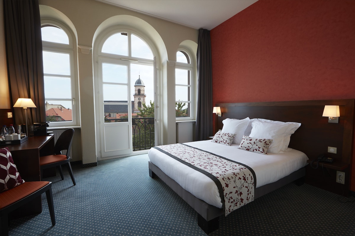 A hotel room with a bed, a desk, a chair, and a view of a building through large arched windows.