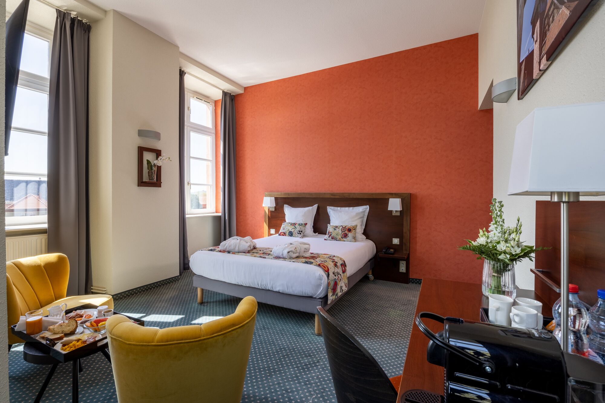 A hotel room with a bed, a yellow armchair, a dining table with breakfast, a nightstand, and a vase with flowers.