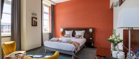 A hotel room with a bed, a yellow armchair, a dining table with breakfast, a nightstand, and a vase with flowers.