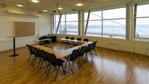 Un salón de conferencias con una larga mesa, sillas, un proyector y grandes ventanales con vistas a un puente y montañas.