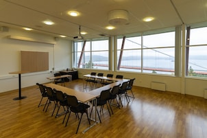Uma sala de conferências com uma mesa comprida, cadeiras, um projetor e janelas amplas com vista para uma ponte e montanhas.