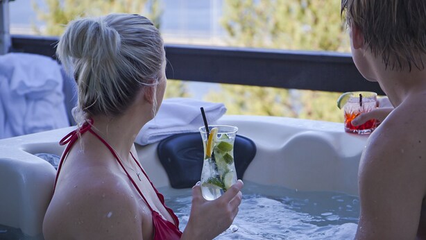 Una persona con traje de baño rojo sosteniendo una bebida en un jacuzzi con vistas a árboles y montañas al fondo.