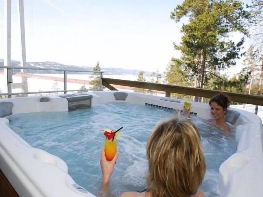 Een bubbelbad met twee personen, waarvan er één een drankje vasthoudt, omringd door sneeuw en bomen.