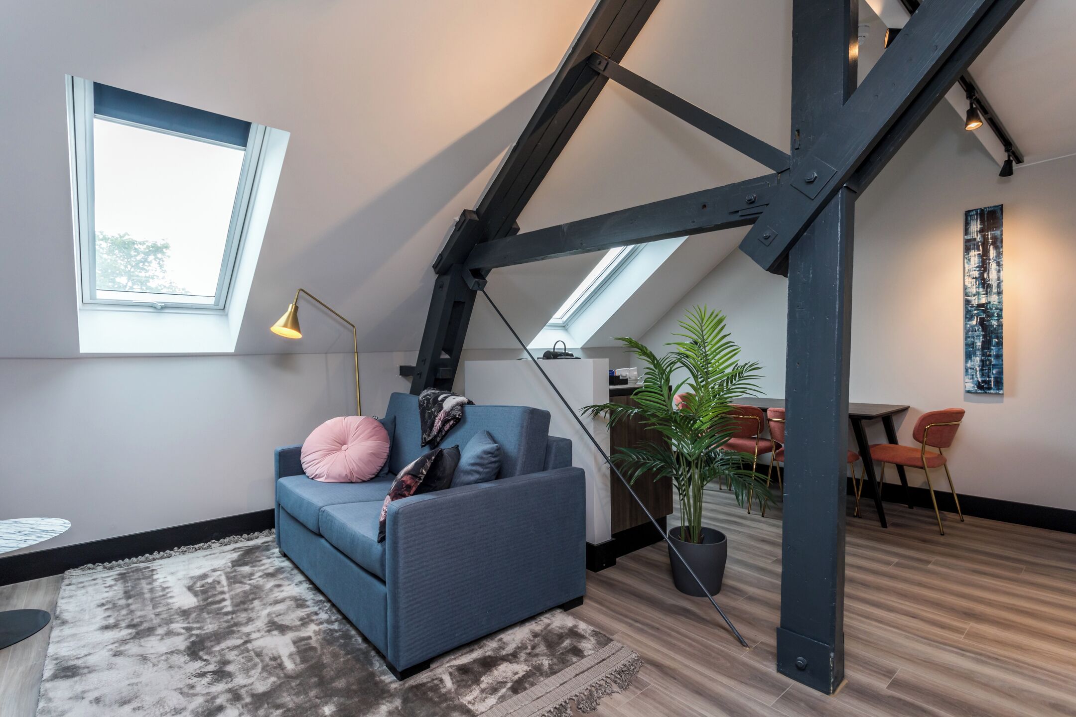 Een loft-achtige kamer met een schuine plafond, een blauwe bank, een klein tafeltje, een vloerlamp, een potplant en een eethoek met stoelen en een tafel.