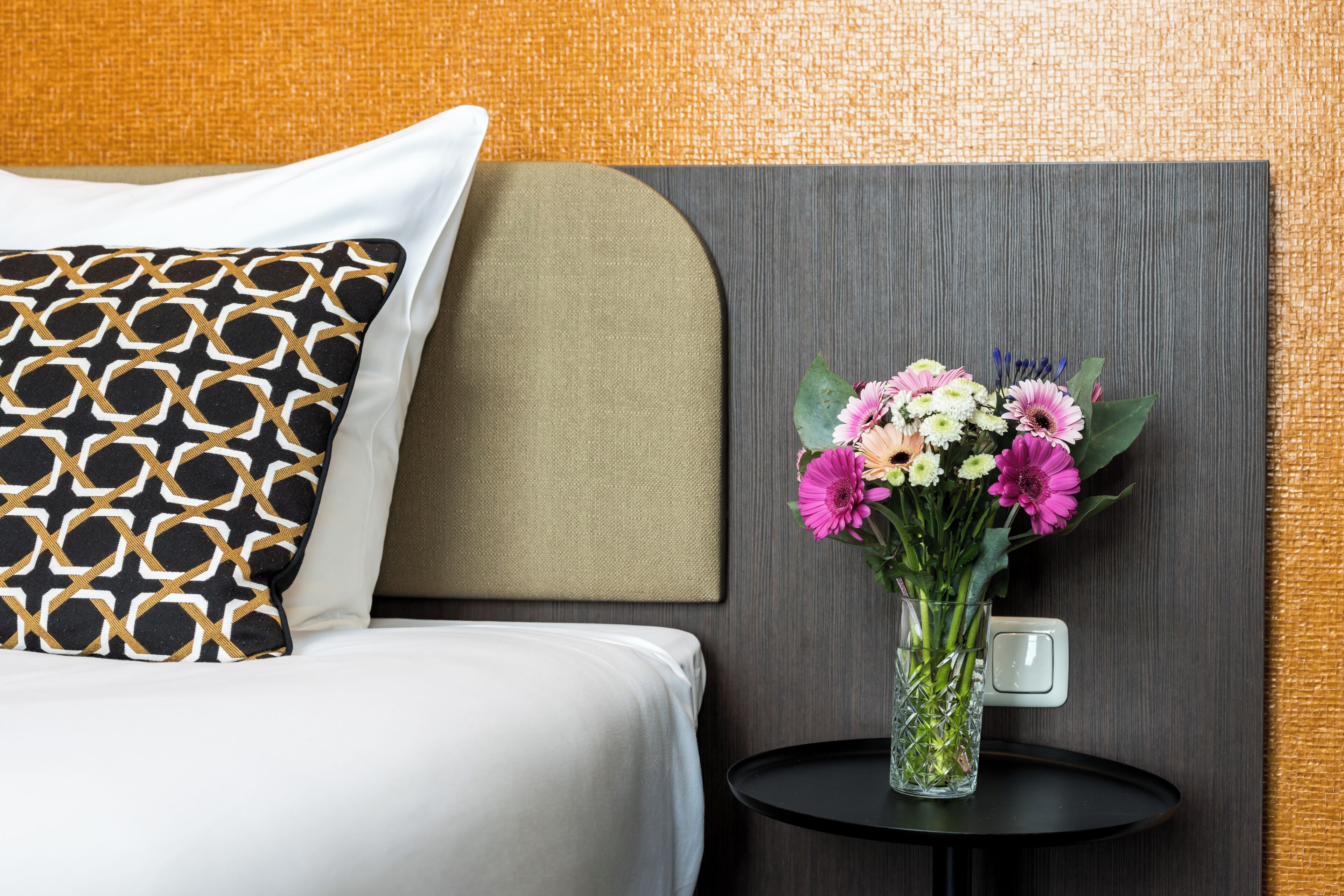 A hotel room with a bed, a decorative pillow, a vase with flowers, and a small table.