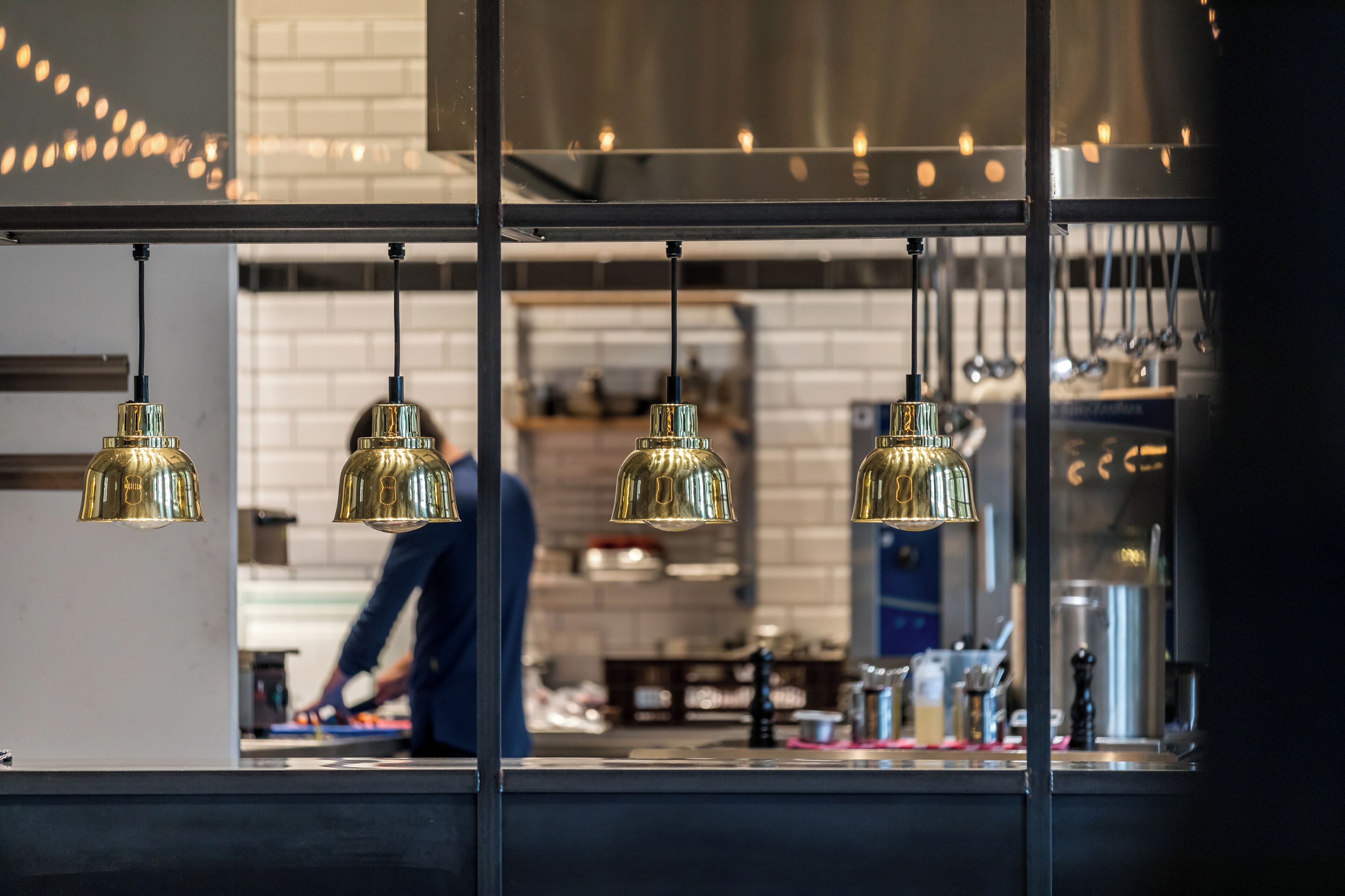 A kitchen with hanging pendant lights, a person working at the counter, and kitchen equipment in the background.