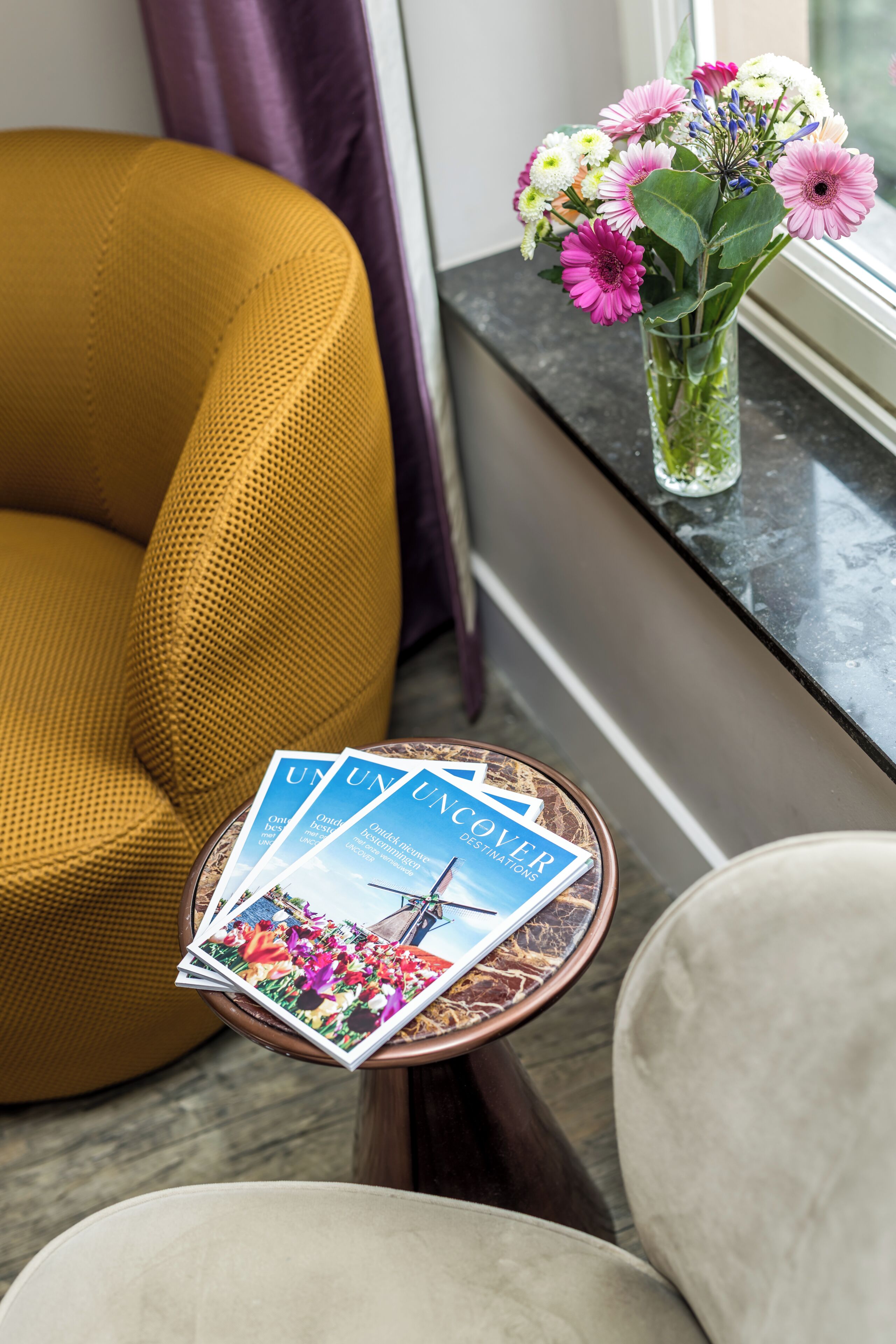 Une pièce avec un fauteuil de couleur moutarde, une petite table sur laquelle repose un magazine et un vase rempli de fleurs près d’une fenêtre.