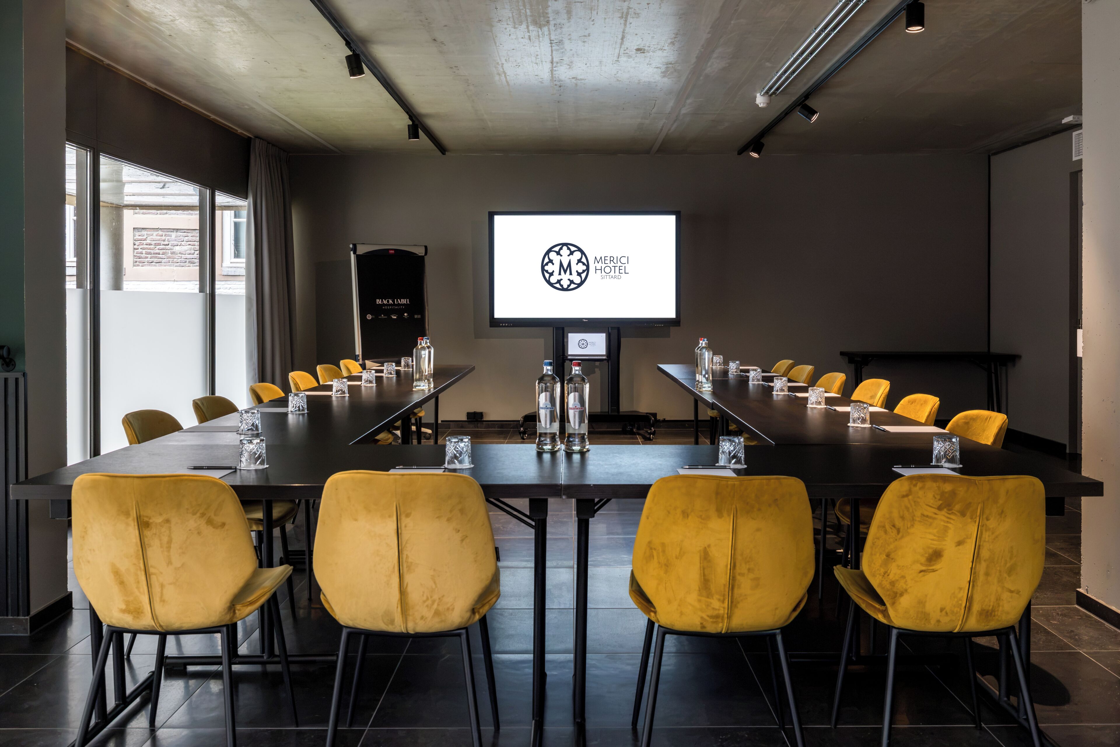 Un salón de conferencias con una larga mesa, sillas amarillas y una pantalla de proyección que muestra un logotipo.