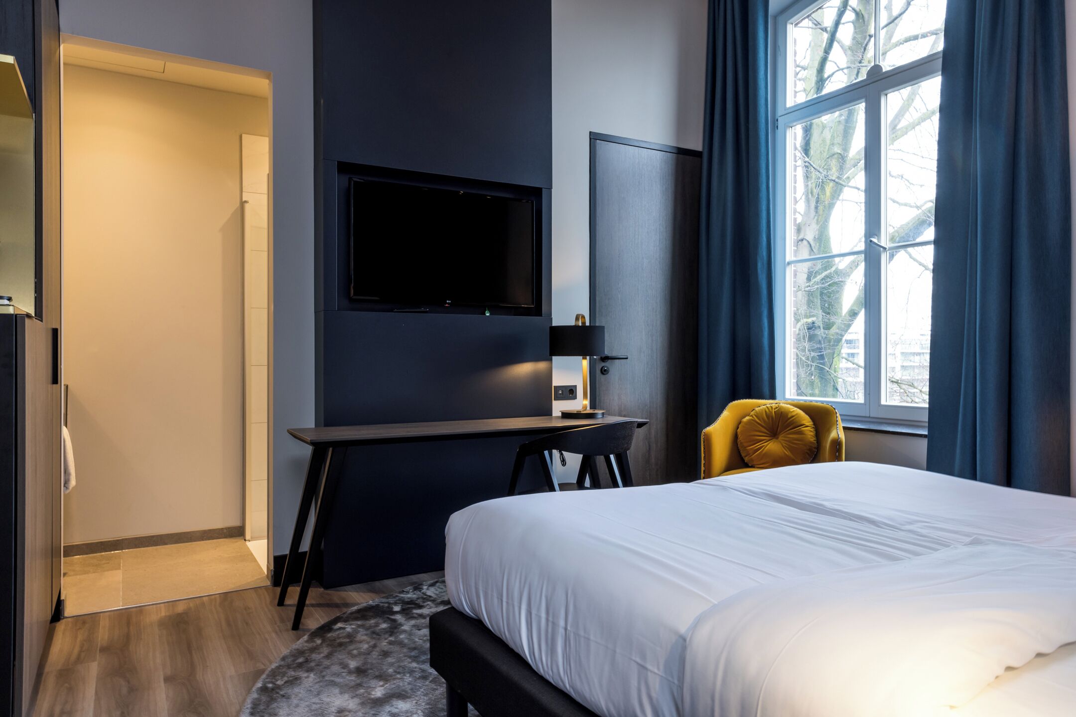 Ett modernt hotellrum med en stor säng, ett skrivbord med en platt-TV, en stol och ett fönster med gardiner.