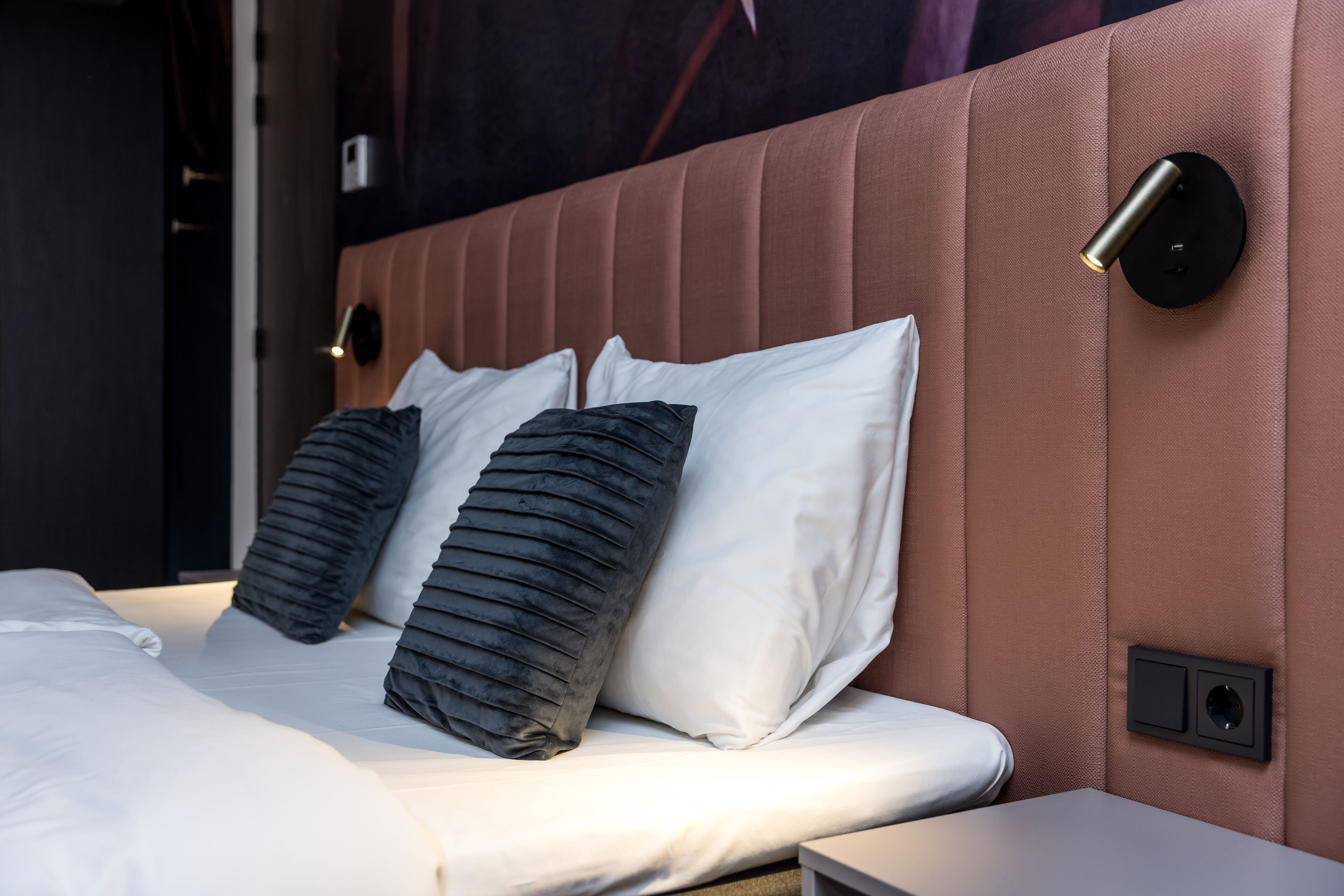 A neatly made bed with white and dark gray pillows, a brown upholstered headboard, and a wall-mounted light fixture.