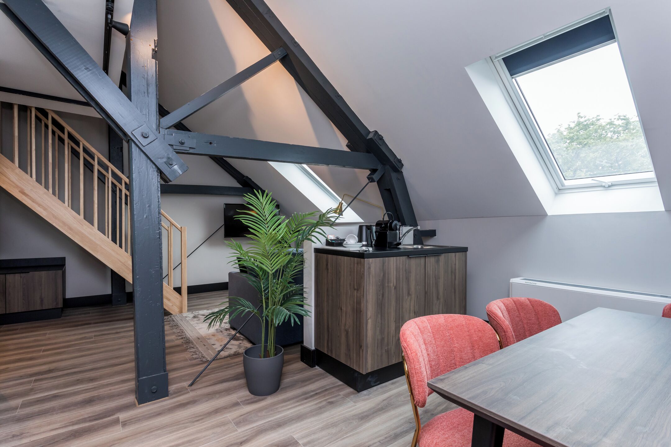 A modern loft-style room with a sloped ceiling, a wooden staircase, a kitchen area with a wooden countertop, and a dining table with red chairs.